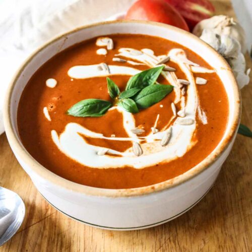 White colour bowl filled with roasted tomato garlic soup topped with cream and basil leaves.