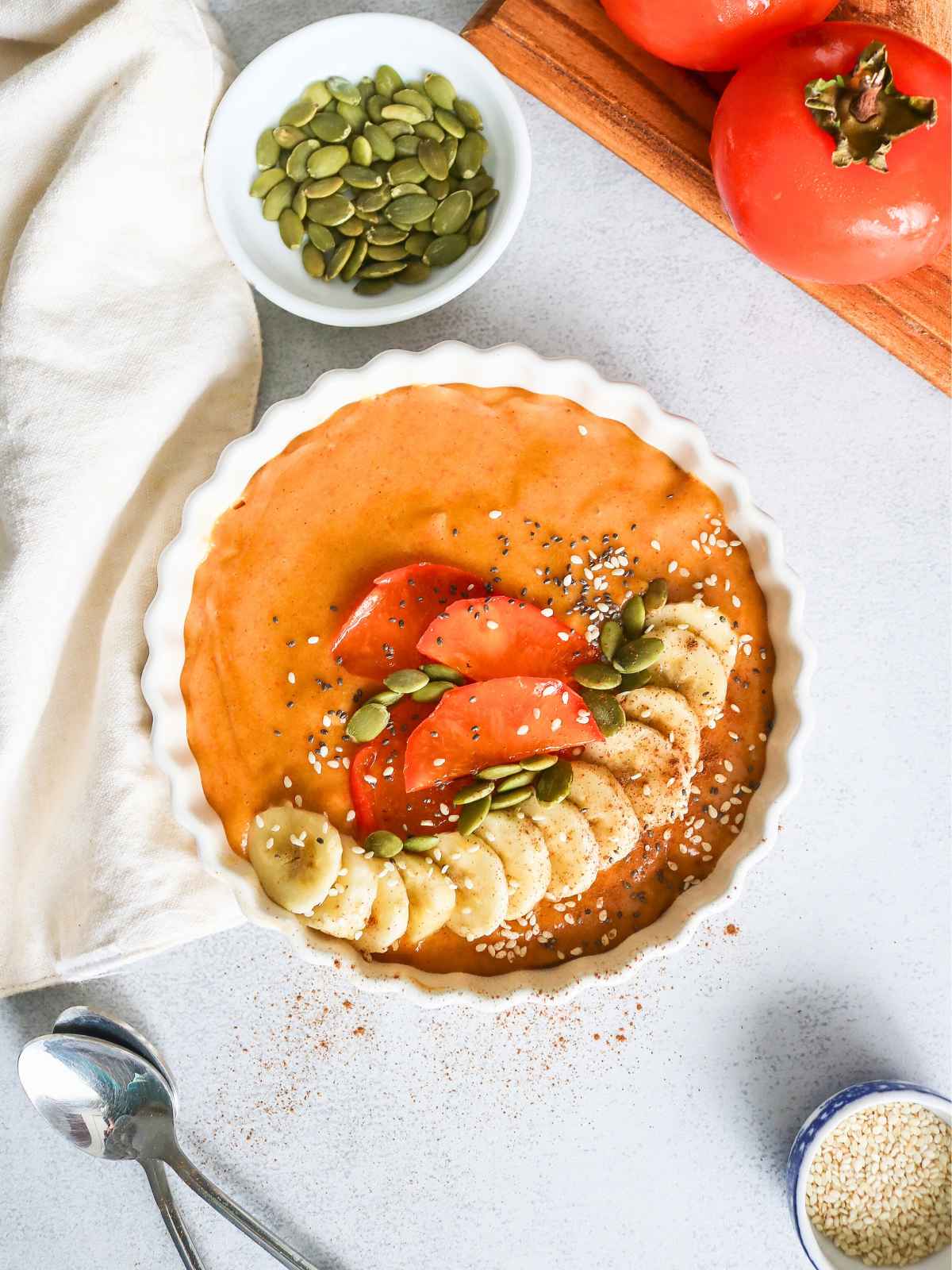 Permission smoothie bowl topped with banana and persimmon slices. Pumpkin seeds on side in a small bowl.