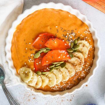 Persimmon smoothie served in a white bowl topped with fresh banana, persimmon slices and pumpkin seeds.
